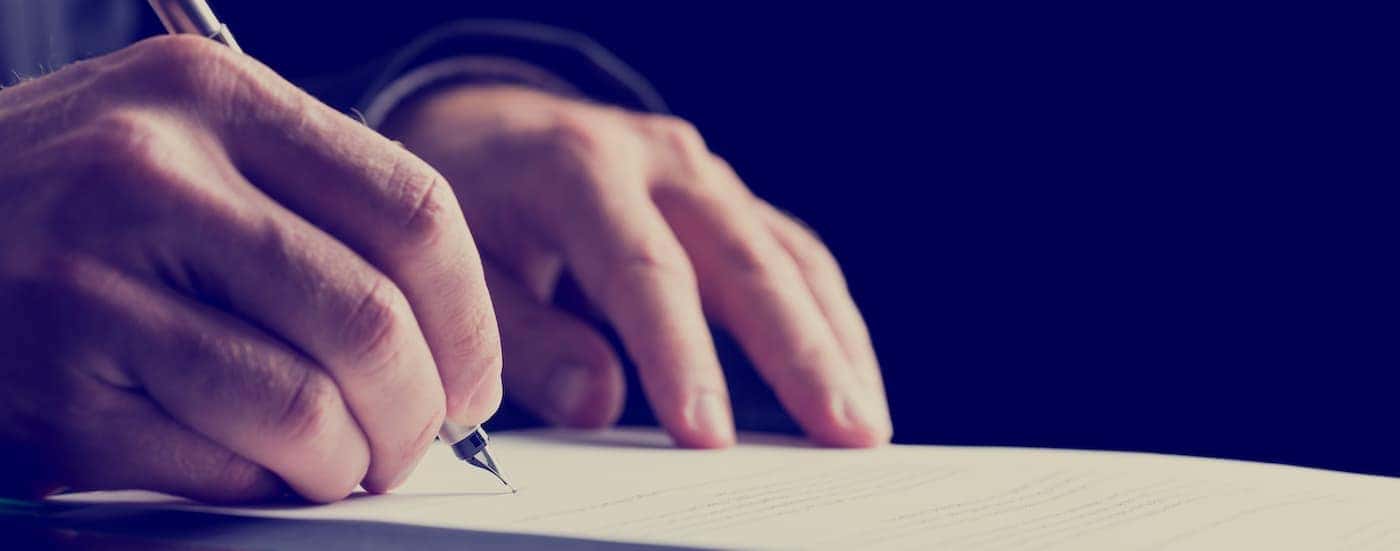 A close up shows a person signing paperwork at a dealership.