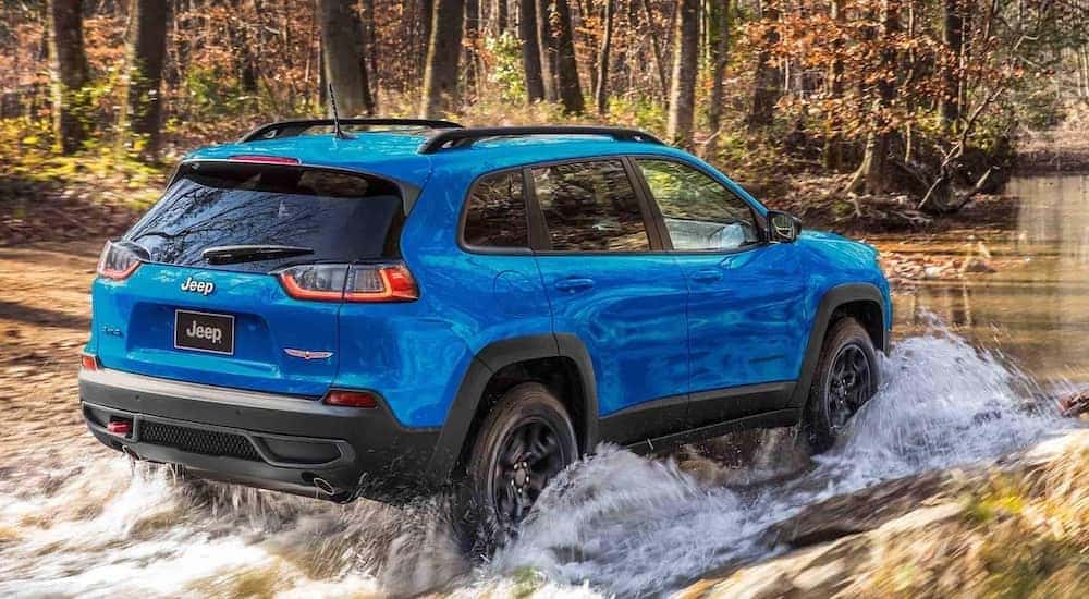 A blue 2023 Jeep Cherokee TrailHawk is shown from the rear at an angle while driving through a river after leaving a dealer that has a Jeep Cherokee for sale.