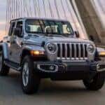 A grey 2021 Jeep Wrangler is shown from the front driving over a bridge.