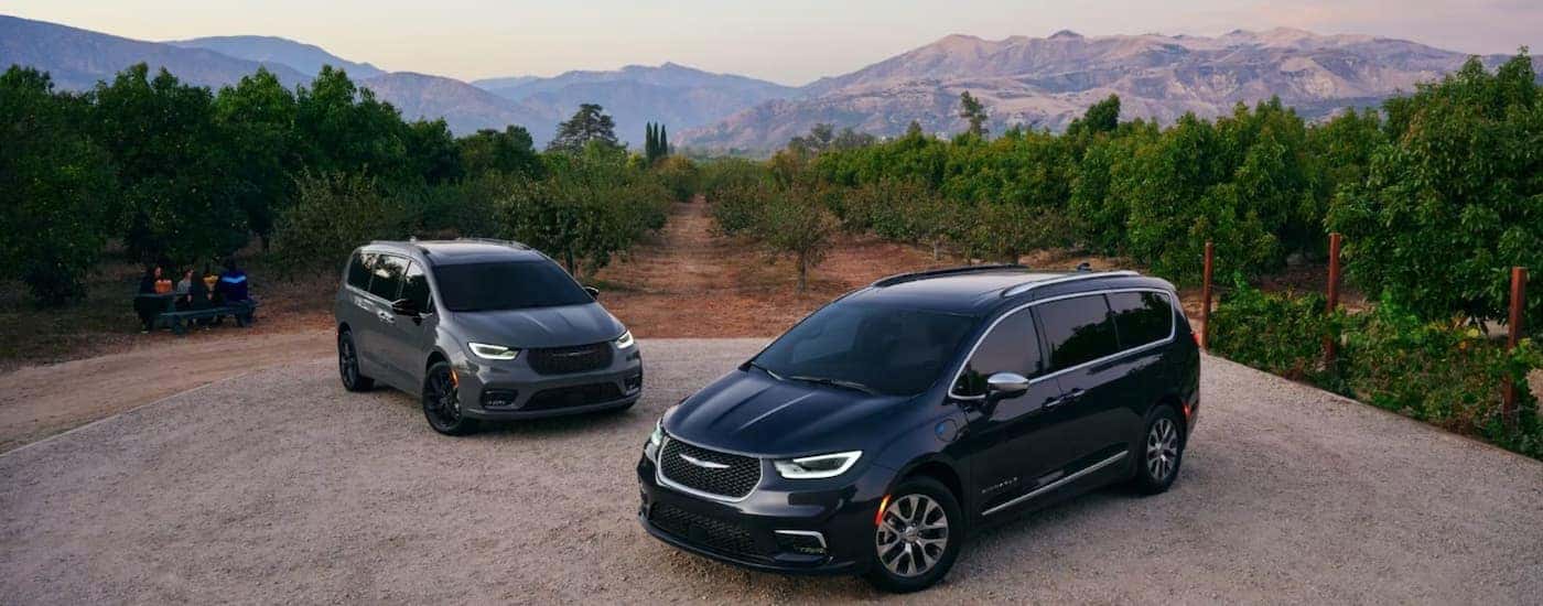 A black and a grey 2023 Chrysler Pacifica are shown parked in a gravel lot with a mountain view.