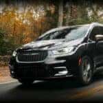 A black 2024 Chrysler Pacifica is shown driving on a tree-lined road.