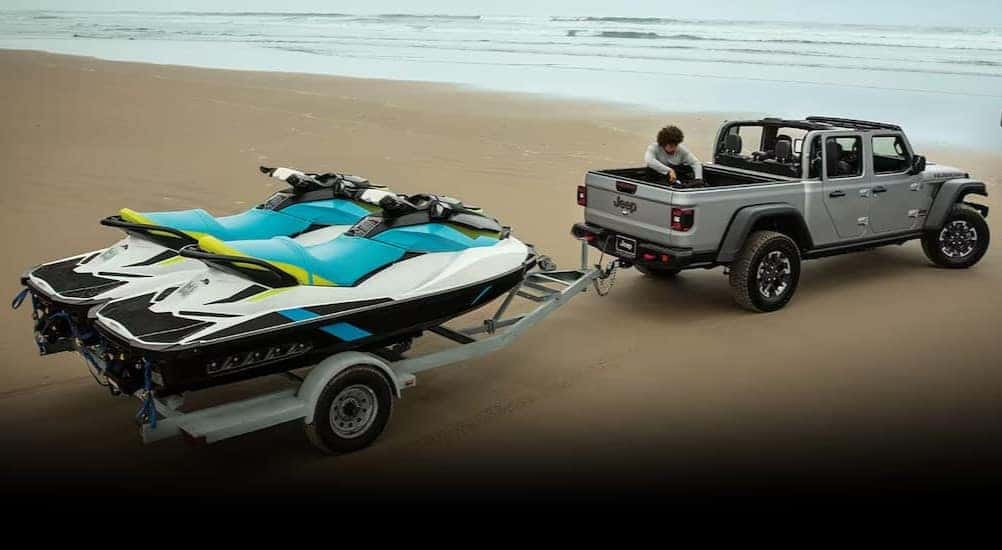A silver 2024 Jeep Gladiator Rubicon is shown parked on a beach while towing two jet skis. 