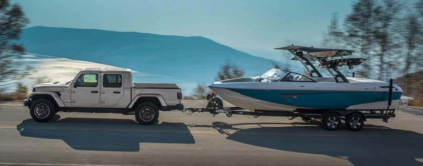 A silver 2023 Jeep Gladiator is towing a boat on a road with a lake view.
