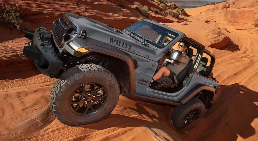 A gray 2025 Jeep Wrangler Willys is shown driving uphill on sand.