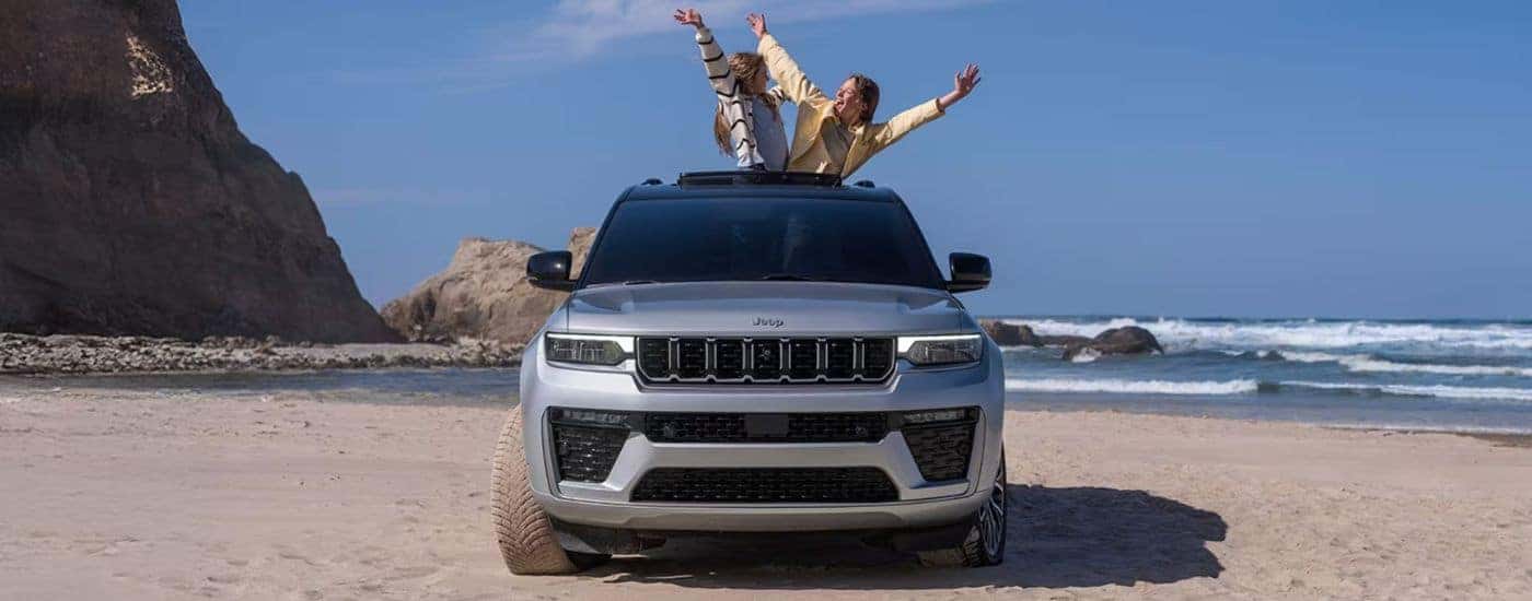 Silver 2026 Jeep Grand Cherokee Summit on the beach with a couple standing out of the sunroof.