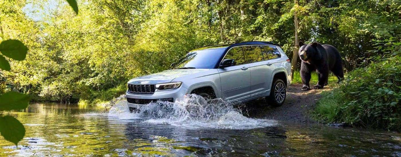 Silver 2026 Jeep Grand Cherokee Summit driving into a stream with a black bear behind it.